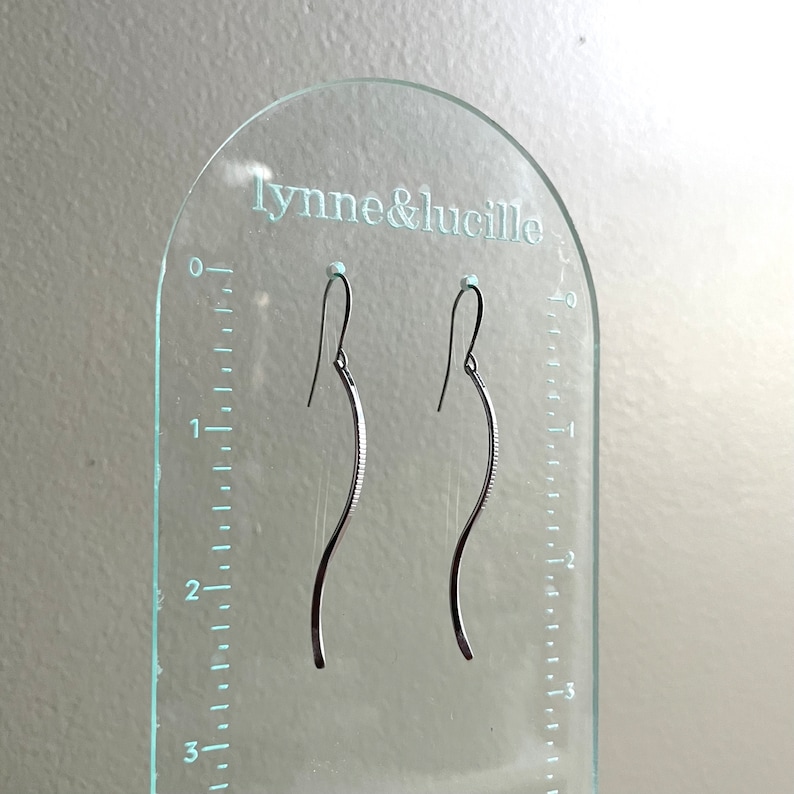May include: A pair of silver-toned, curved earrings with a textured design. The earrings are displayed on a clear acrylic stand with the text "lynne&lucille". The stand has a ruler printed on the side.