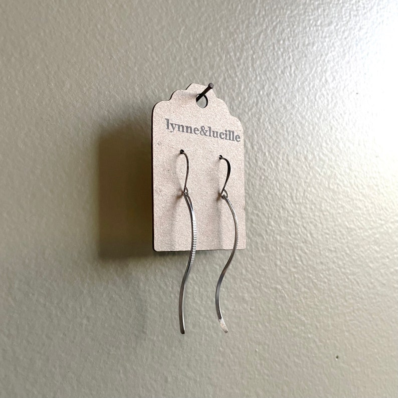 May include: Silver-toned, wavy dangle earrings hanging from a cardboard tag. The earrings have a simple, elegant design with a textured, linear pattern on one side. The tag is labeled "lynne&lucille".