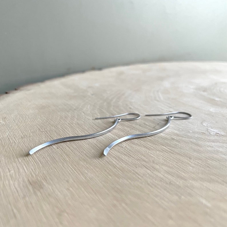 May include: A pair of silver-toned, curved dangle earrings. The earrings have a simple, minimalist design with a slender, wavy shape. The hooks are visible at the top, and the earrings are displayed on a light-colored wooden surface.