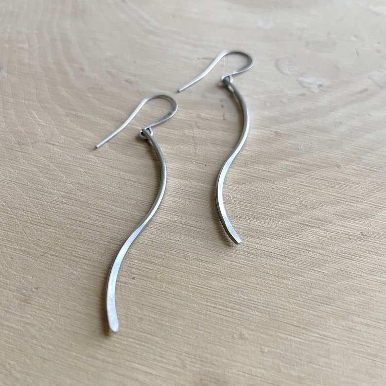 May include: A pair of silver-toned, curved dangle earrings. Each earring features a slender, wavy design with a hook for pierced ears. The earrings are displayed on a light-colored wooden surface.