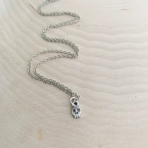 May include: A silver-colored necklace with a small skull pendant. The pendant has two eye sockets and a nose cavity. The chain is delicate and hangs on a light-colored wooden surface.