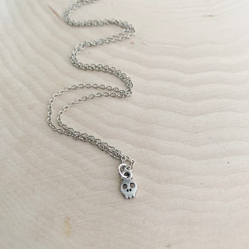 May include: A silver-colored necklace with a small skull pendant. The pendant has two eye sockets and a nose cavity. The chain is delicate and hangs on a light-colored wooden surface.