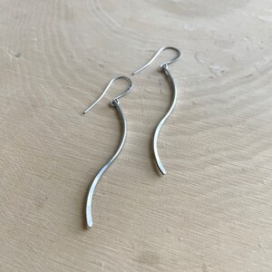 May include: A pair of silver-toned, curved dangle earrings. The earrings have a simple, flowing design with a gentle wave shape. The hooks are visible at the top, and the earrings are displayed on a light-colored wooden surface.