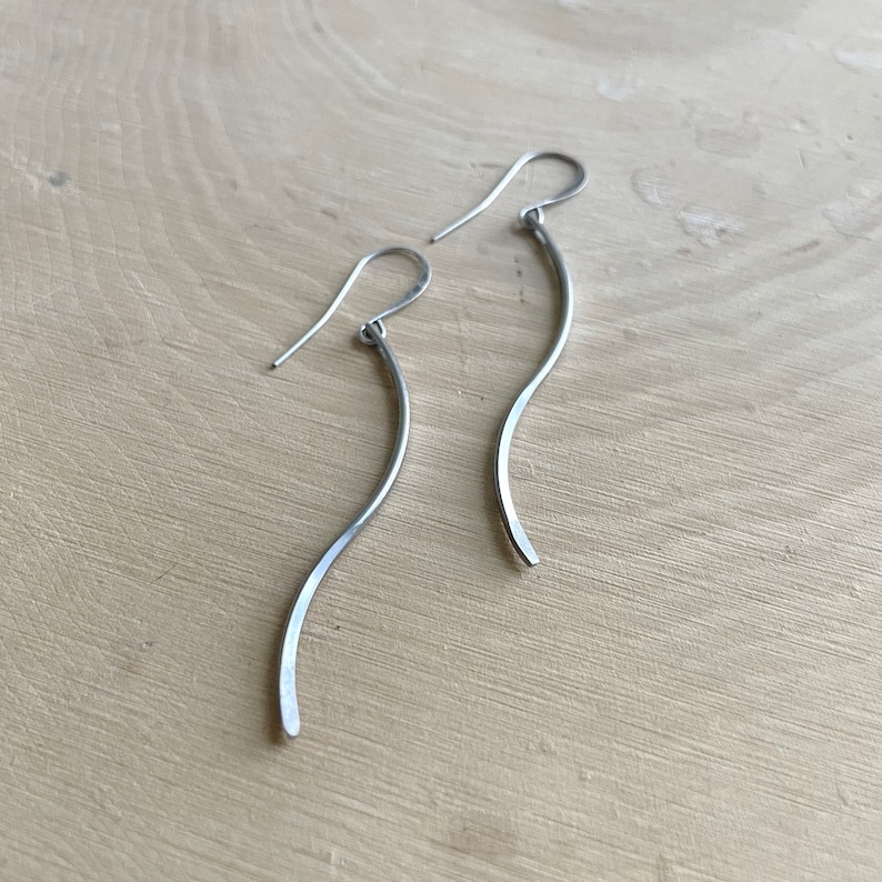 May include: A pair of silver-toned, curved dangle earrings. The earrings have a simple, flowing design with a gentle wave shape. The hooks are visible at the top, and the earrings are displayed on a light-colored wooden surface.