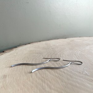 May include: A pair of silver-toned, wavy dangle earrings. The earrings have a simple, minimalist design with a textured surface. They are displayed on a light-colored wooden surface.