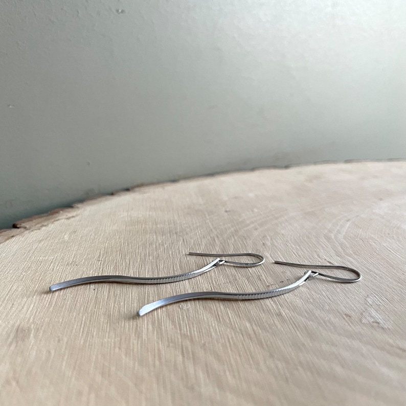May include: A pair of silver-toned, wavy dangle earrings. The earrings have a simple, minimalist design with a textured surface. They are displayed on a light-colored wooden surface.