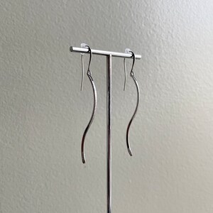 May include: A pair of silver-toned, curved dangle earrings. The earrings have a textured, ridged design on the inner curve and a smooth, polished finish on the outer curve. They are displayed on a silver earring stand.