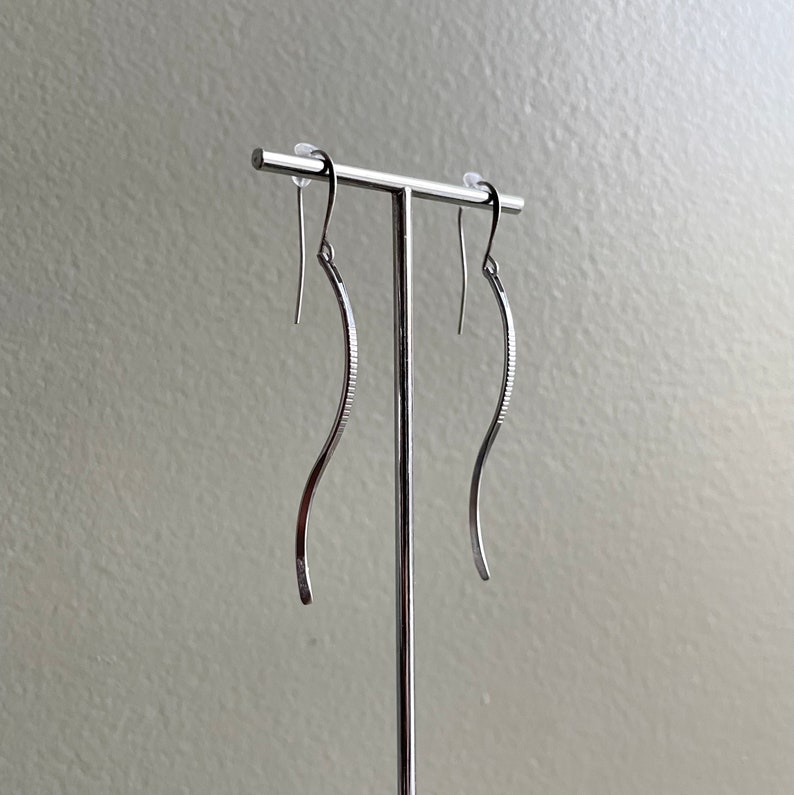 May include: A pair of silver-colored, curved dangle earrings. The earrings have a slender, wavy design with a textured, ridged detail on the inner curve. They are displayed on a silver earring stand against a neutral background.