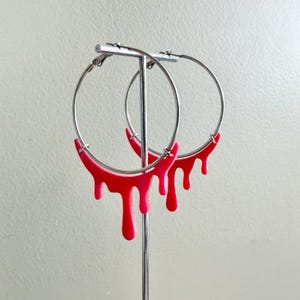May include: A pair of silver hoop earrings with a unique design. The bottom half of each earring is a vibrant red, with a dripping effect. The earrings are displayed on a silver earring stand against a neutral background.