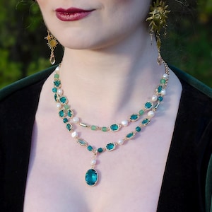 Op de afbeelding: Een vrouw draagt een groene fluwelen top en een gelaagde ketting met smaragdgroene en parelaccenten. De ketting heeft een grote, druppelvormige teal-kleurige hanger. Goudkleurige sterrenoorbellen maken de look compleet. De tekst "Ithuriel handcrafted jewellery" is zichtbaar.