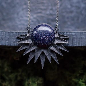 May include: A dark, celestial-themed necklace featuring a blue-speckled orb pendant set within a spiky, starburst-shaped frame. The pendant hangs from a delicate silver chain. The brand name "Ithuriel" is visible at the bottom.