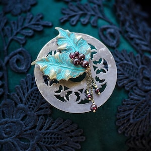May include: A handcrafted brooch with a teal and gold-toned holly leaf design and red berry cluster. The brooch sits on a silver-coloured, circular base with cut-out patterns. A chain with red beads adds detail. The brand name 'Ithuriel' is visible.