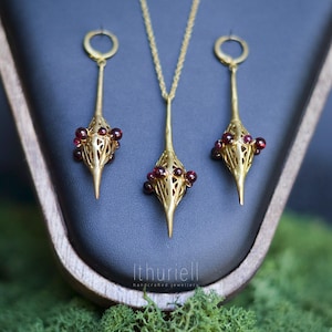 May include: A gold-toned jewellery set comprising a pendant necklace and matching earrings. Each piece features an elongated design with a cage-like structure, embellished with small, red gemstones. The jewellery is presented on a dark blue surface.