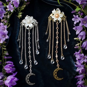 May include: Two hair accessories featuring a white flower with a central pearl, dangling silver and gold chains, clear teardrop-shaped crystals, and a crescent moon charm at the base. The text Ithuriel handcrafted jewellery is at the bottom.