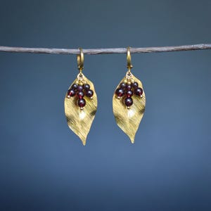 May include: A pair of gold-coloured leaf earrings with a textured finish, each featuring a cluster of small, dark red beads. The earrings hang from gold hoops against a blurred blue backdrop. The brand name "lithuriell" is visible at the bottom.