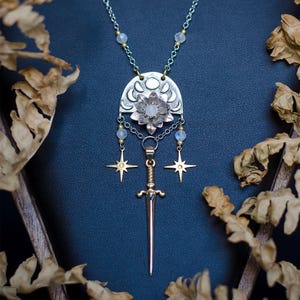 May include: A silver necklace with a floral pendant and a small sword charm. The pendant features a flower design with a moonstone centre and is accented with gold starburst charms. The necklace is displayed on a dark blue surface.