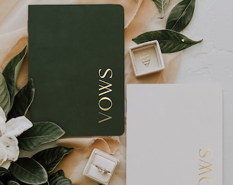 May include: Two white notebooks with the word "Vows" written in gold lettering on each. The notebooks are surrounded by green leaves, a white flower, and a beige fabric. Two small white boxes with rings inside are also visible.