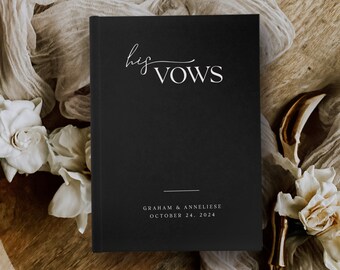 May include: Black vow book with white text that reads "his vows" and "Graham & Anneliese October 24, 2024". The book is on a wooden surface with white fabric and flowers.