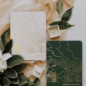 May include: Two wedding invitations with a topographic map design. The invitations are on white and dark green cardstock with gold foil accents. The text on the invitations reads "Ashton & Josephine August 18, 2024".