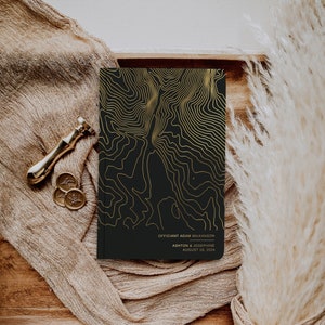 May include: A black wedding ceremony programme with gold foil accents and a topographic design. The text reads "Officiant Adam Wilkinson" and "Ashton & Josephine August 18, 2023".