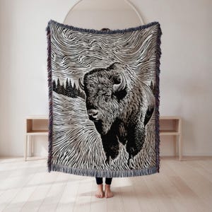 May include: A black and white woven throw blanket featuring a detailed illustration of a bison in a field. The blanket has a fringed edge with a mix of colors. The design includes a landscape with trees and a textured sky.