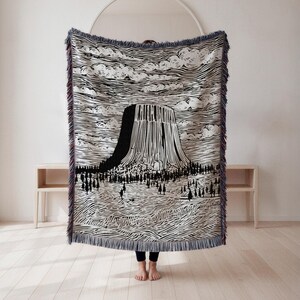May include: A black and white woven throw blanket featuring a graphic of Devils Tower National Monument. The design includes a detailed illustration of the tower, clouds, and a forest. The blanket has fringed edges and is displayed in a room with light-colored wood flooring.