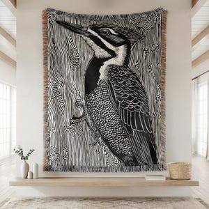 May include: A large black and white woven throw blanket featuring a detailed illustration of a woodpecker. The bird is set against a background of vertical wood grain lines. The blanket has a fringed edge.