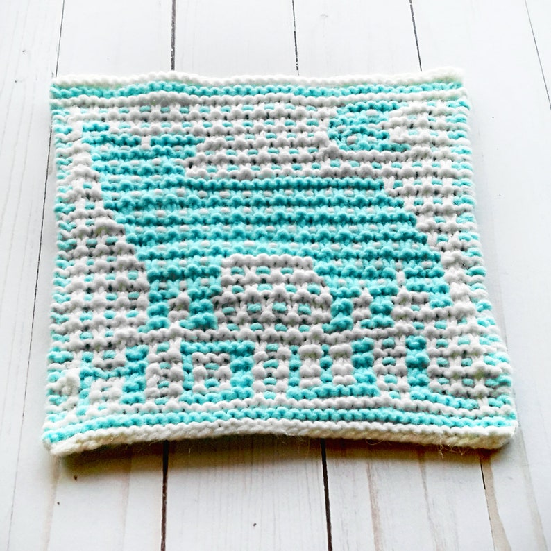 May include: A knitted dishcloth in a blue and white colour pattern. The dishcloth is square shaped and has a textured surface.