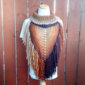 Pode incluir: Um poncho de malha com um padr&atilde;o degrad&ecirc; marrom, laranja e roxo e franjas. O poncho tem um pesco&ccedil;o alto e &eacute; feito de um fio macio e quente.