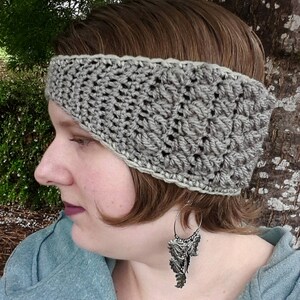 May include: A grey crocheted headband with a white border. The headband is worn by a person with brown hair and is angled to cover one ear.
