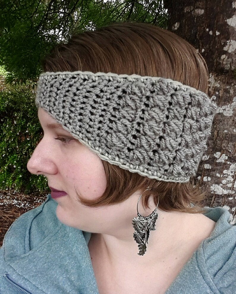 May include: A grey crocheted headband with a white border. The headband is worn by a person with brown hair and is angled to cover one ear.