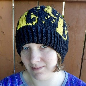 Op de afbeelding: Een zwarte gebreide beanie met een geel gebreid ontwerp. De beanie heeft een geribbelde textuur en een omgeslagen rand.