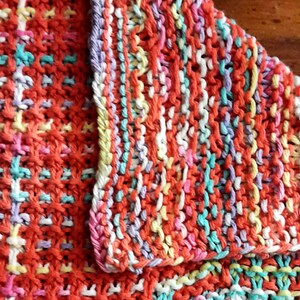 May include: A close-up of a knitted dishcloth with a colorful striped pattern in shades of orange, pink, white, yellow, and blue.