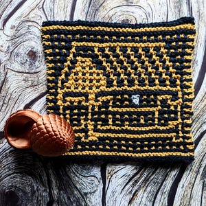 May include: A black and gold crocheted square with a geometric pattern, and a small, copper-colored acorn-shaped object. The square has a detailed design, and the acorn-shaped object is next to the square.