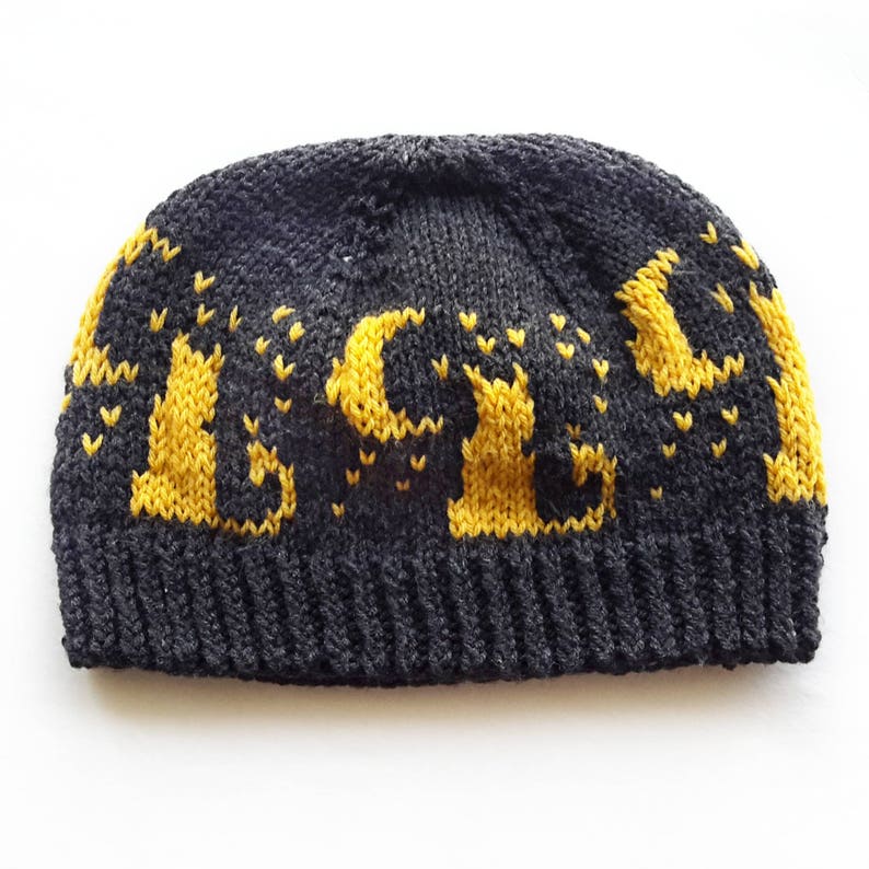 Op de afbeelding: Een zwarte gebreide beanie met een geel gebreid ontwerp van het woord "ALIVE".