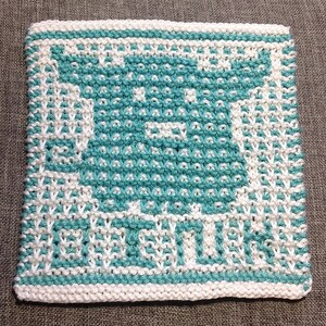 K&ouml;nnte beinhalten: Ein wei&szlig;er gestrickter Geschirrtuch mit einem blaugr&uuml;nen Design einer Teetasse und den Worten "tea time".