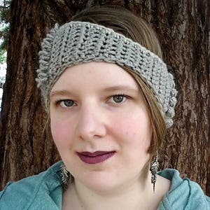 May include: A woman wearing a grey crocheted headband with a textured design. She is wearing a teal blue shirt and silver earrings.