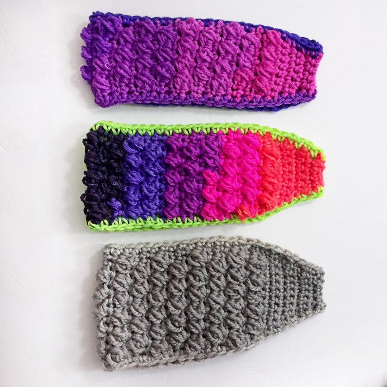 May include: Three crocheted headbands in different colours. The top headband is purple and pink, the middle headband is black, purple, pink, and green, and the bottom headband is grey.