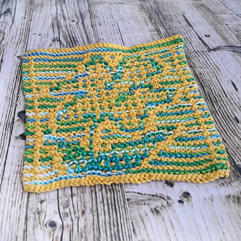 May include: A yellow knitted dishcloth with a green, blue and white pattern. The dishcloth is made with a simple knit stitch and has a textured surface.