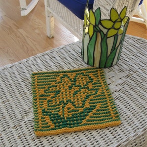 May include: A yellow and green knitted dishcloth with a floral design. The dishcloth is sitting on a white wicker table with a stained glass lampshade featuring yellow flowers.