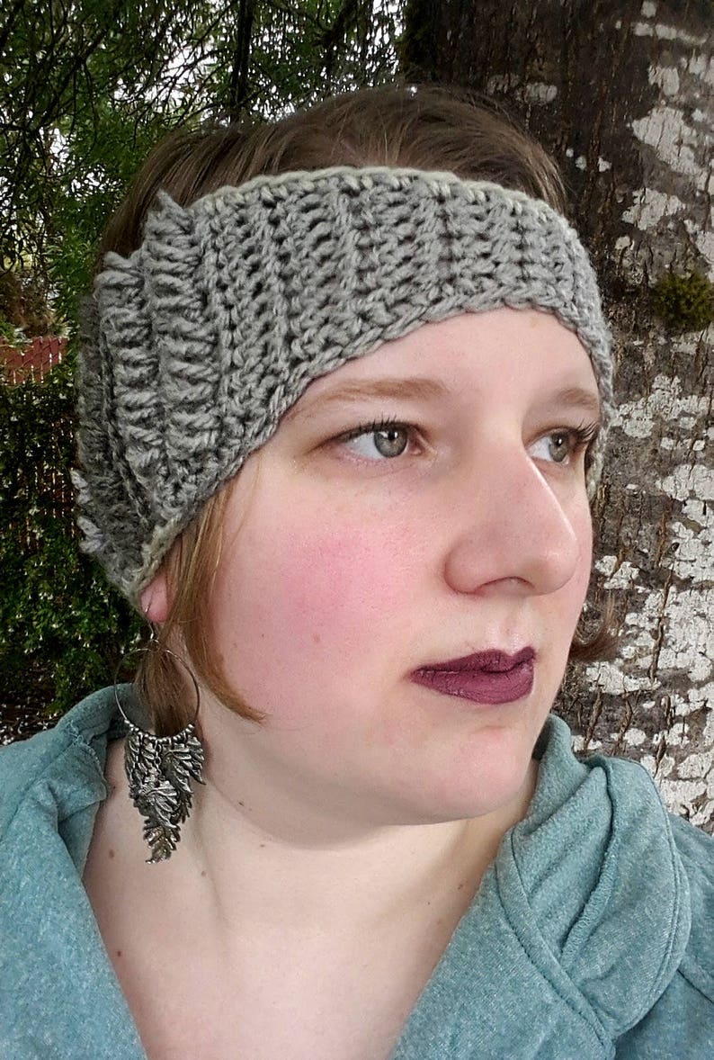 May include: A grey crocheted headband with a white trim. The headband is being worn by a person with brown hair and a light blue hooded sweatshirt. The person is wearing silver hoop earrings with a dangling feather design.