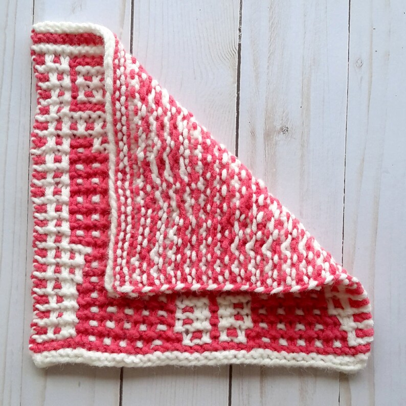 May include: A pink and white knitted dishcloth with a textured pattern. The dishcloth is folded in half and the edges are visible.