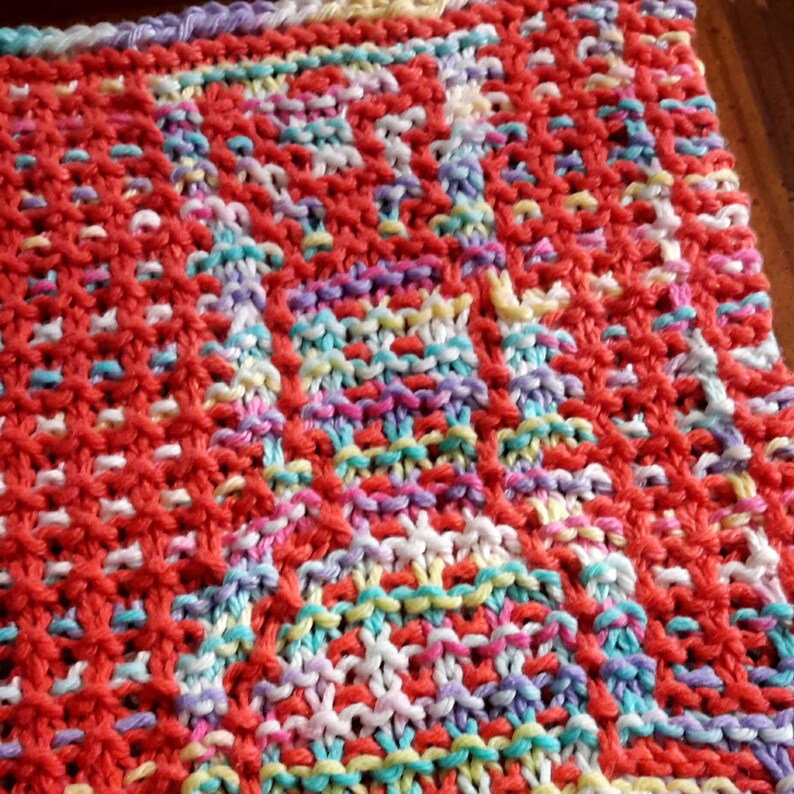 May include: Close-up of a knitted dishcloth with a colorful, multi-colored pattern. The dishcloth is made with a variety of yarn colors, including red, pink, white, blue, and yellow.
