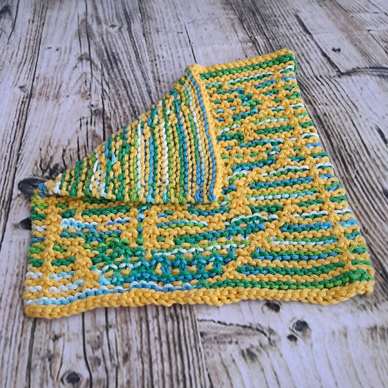 May include: A yellow knitted dishcloth with green, blue and white stripes. The dishcloth is folded in half and is lying on a wooden surface.