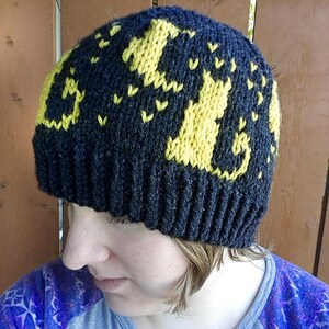 Op de afbeelding: Zwarte gebreide beanie met een geel gebreid ontwerp van katten. De beanie heeft een geribbelde rand.
