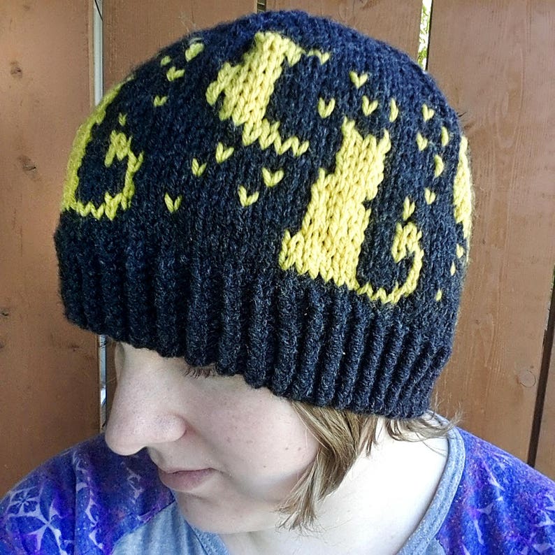 Op de afbeelding: Zwarte gebreide beanie met een geel gebreid ontwerp van katten. De beanie heeft een geribbelde rand.