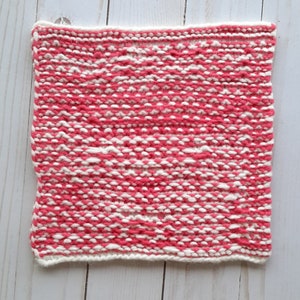 May include: A pink and white knitted square with a textured pattern. The square is made with a soft yarn and has a white border.