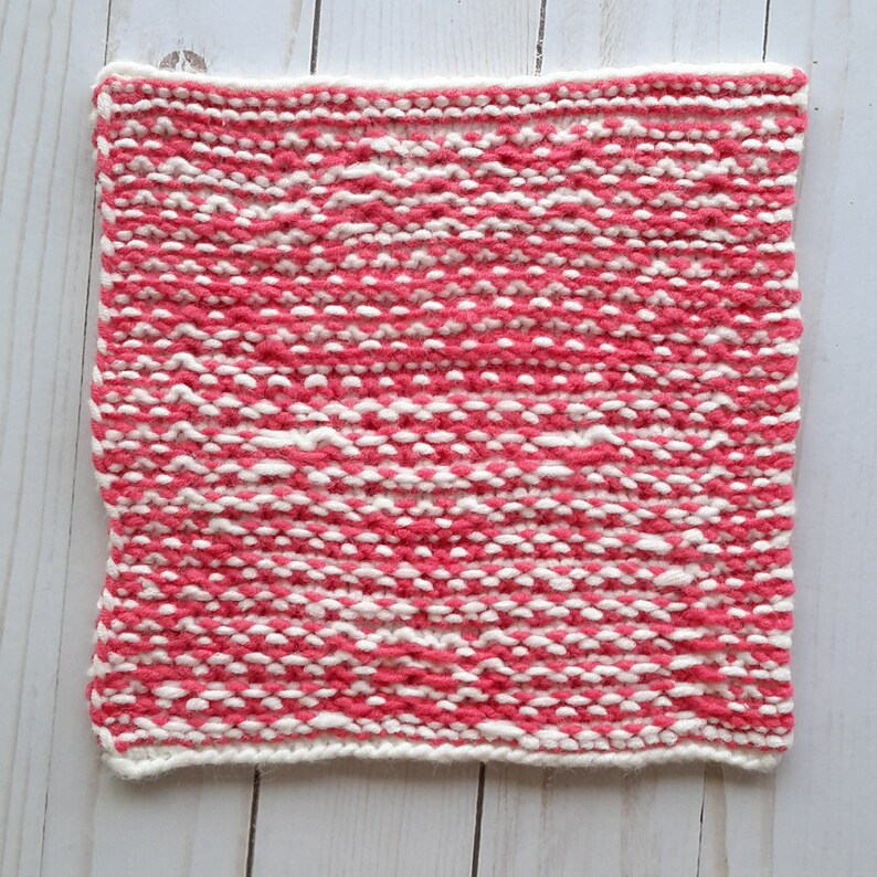 May include: A pink and white knitted square with a textured pattern. The square is made with a soft yarn and has a white border.