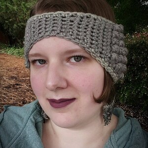 May include: A woman wearing a grey crocheted headband with a textured design. The headband covers part of her forehead and one ear. She is wearing a grey sweater and silver earrings.
