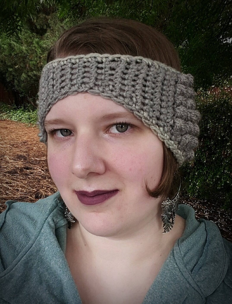 May include: A woman wearing a grey crocheted headband with a textured design. The headband covers part of her forehead and one ear. She is wearing a grey sweater and silver earrings.